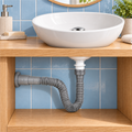 Sifón flexible gris con push cromado instalado bajo lavamanos, conectado al desagüe del muro
